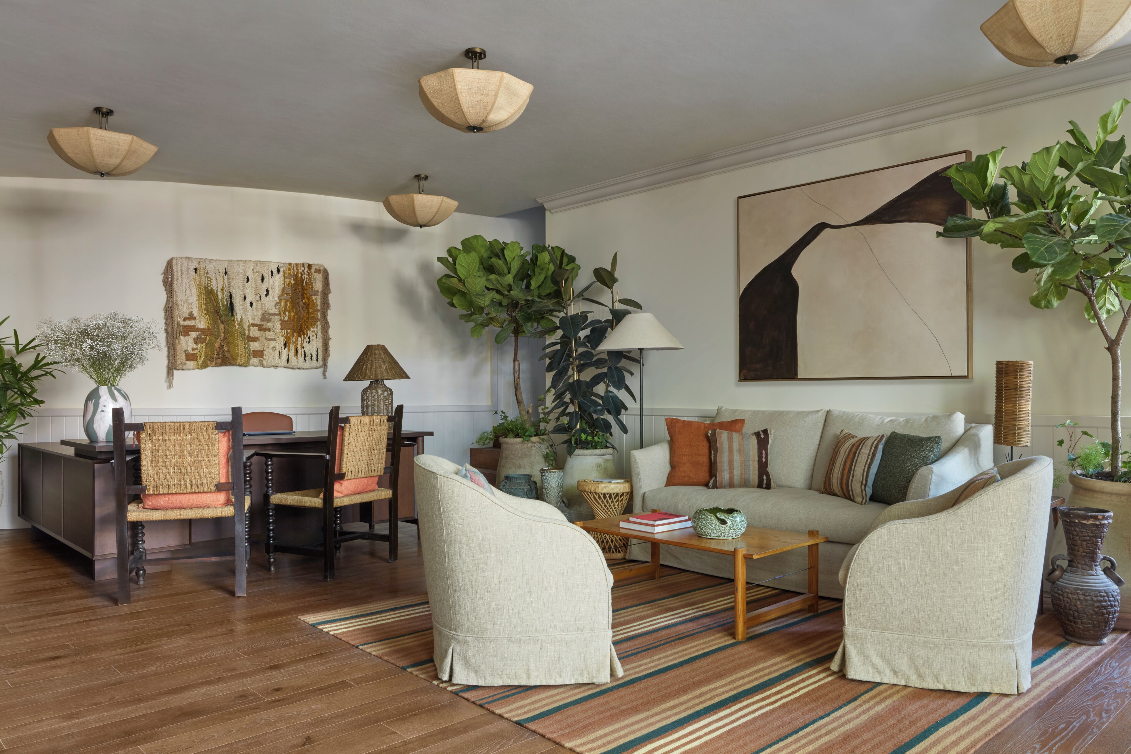 A living room with a sofa, chairs, a coffee table, and a dining table with chairs. There are plants, a lamp, and artwork on the walls.