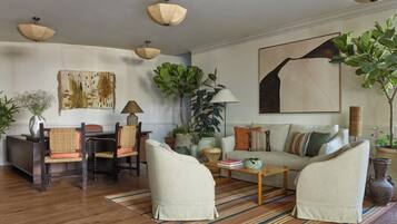 Ein Wohnzimmer mit einer Couch, Sesseln, einem Couchtisch und einem Esstisch mit Stühlen. Es gibt Pflanzen, eine Lampe und Kunstwerke an den Wänden.