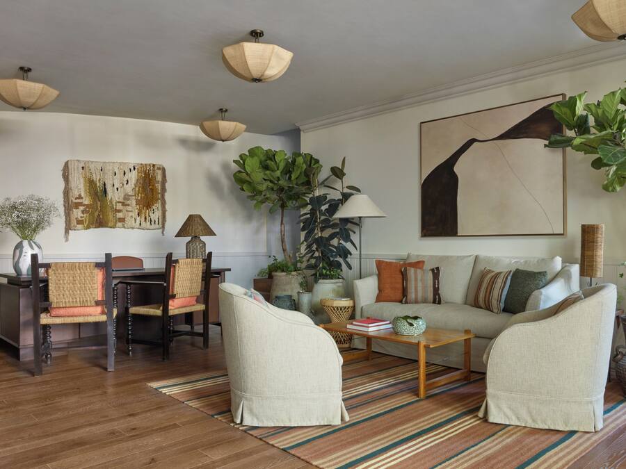 Ein Wohnzimmer mit einer Couch, Sesseln, einem Couchtisch und einem Esstisch mit Stühlen. Es gibt Pflanzen, eine Lampe und Kunstwerke an den Wänden.