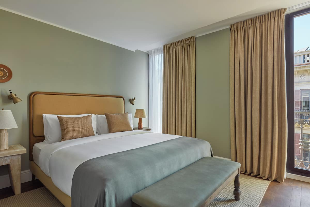A hotel room with a large bed, a bench, two bedside lamps, a window with curtains, and a view of the outside.