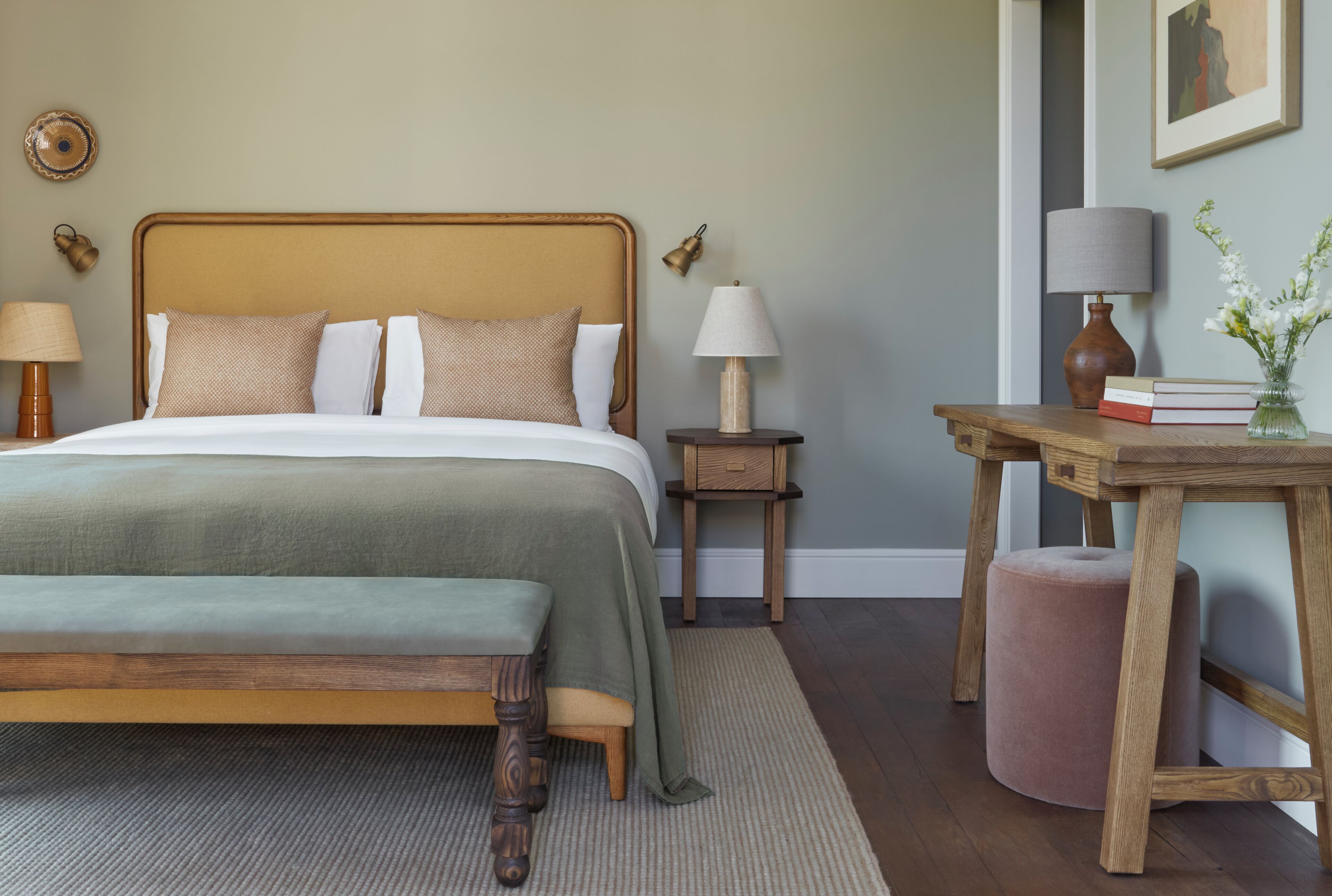 Una cama bien hecha con cabecera de madera, una mesita de noche con lámpara, un taburete pequeño de madera y un florero con flores.