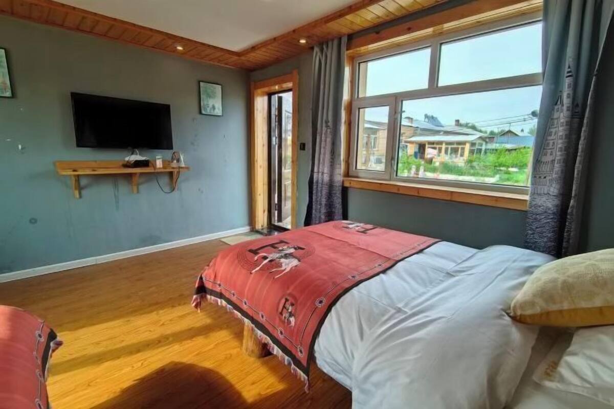 Een slaapkamer met een bed, een aan de muur bevestigde televisie, een raam met gordijnen en een houten vloer.