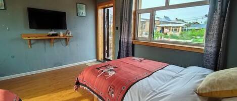 Un dormitorio con una cama, un televisor montado en la pared, una ventana con vistas y suelo de madera.