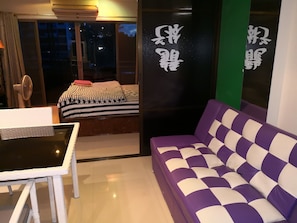 Une pièce comprenant un lit, un canapé violet et blanc, une table et une chaise. Il y a une fenêtre avec des rideaux et une vue sur l’extérieur.