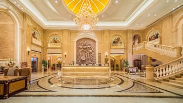A grand lobby with a central reception desk, a large chandelier, and a grand staircase.