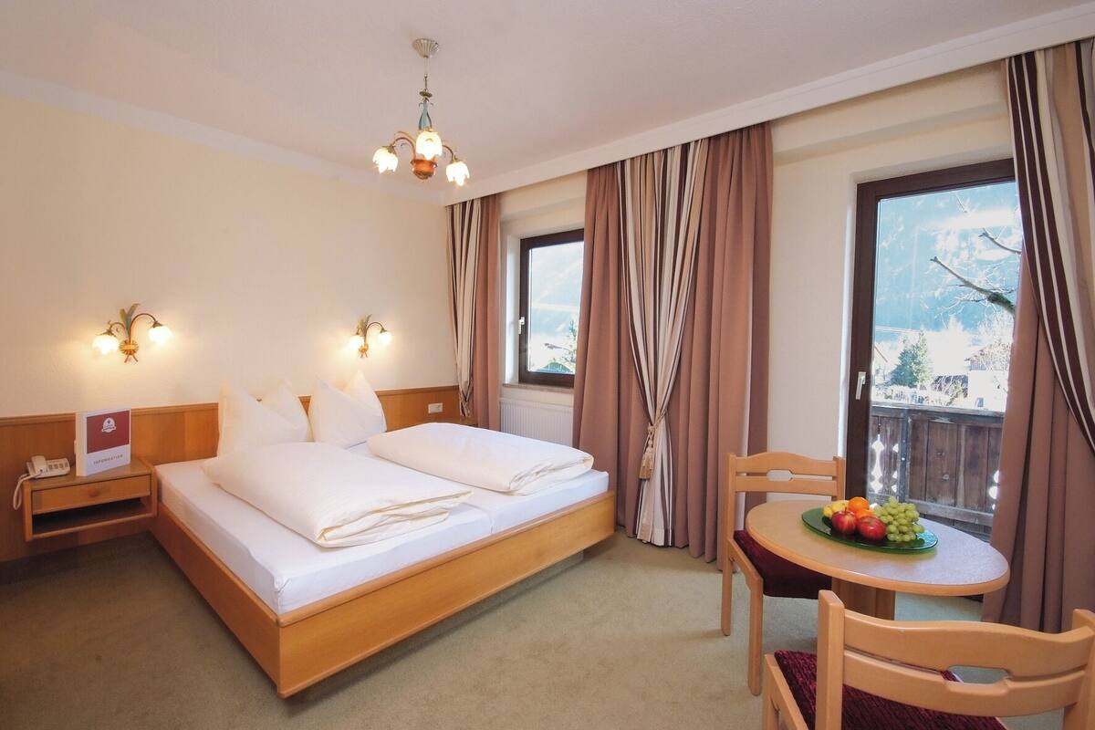 A hotel room with a bed, bedside table with a phone, a table with fruit, and a view of the outdoors.