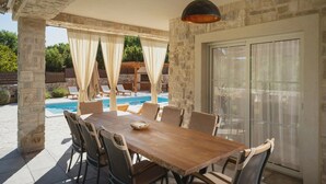 An outdoor patio with a wooden table and chairs, a stone wall, and a view of a swimming pool. - Lulu in Dobrinj With 4 Bedrooms and 4 Bathrooms (Dobrinj)