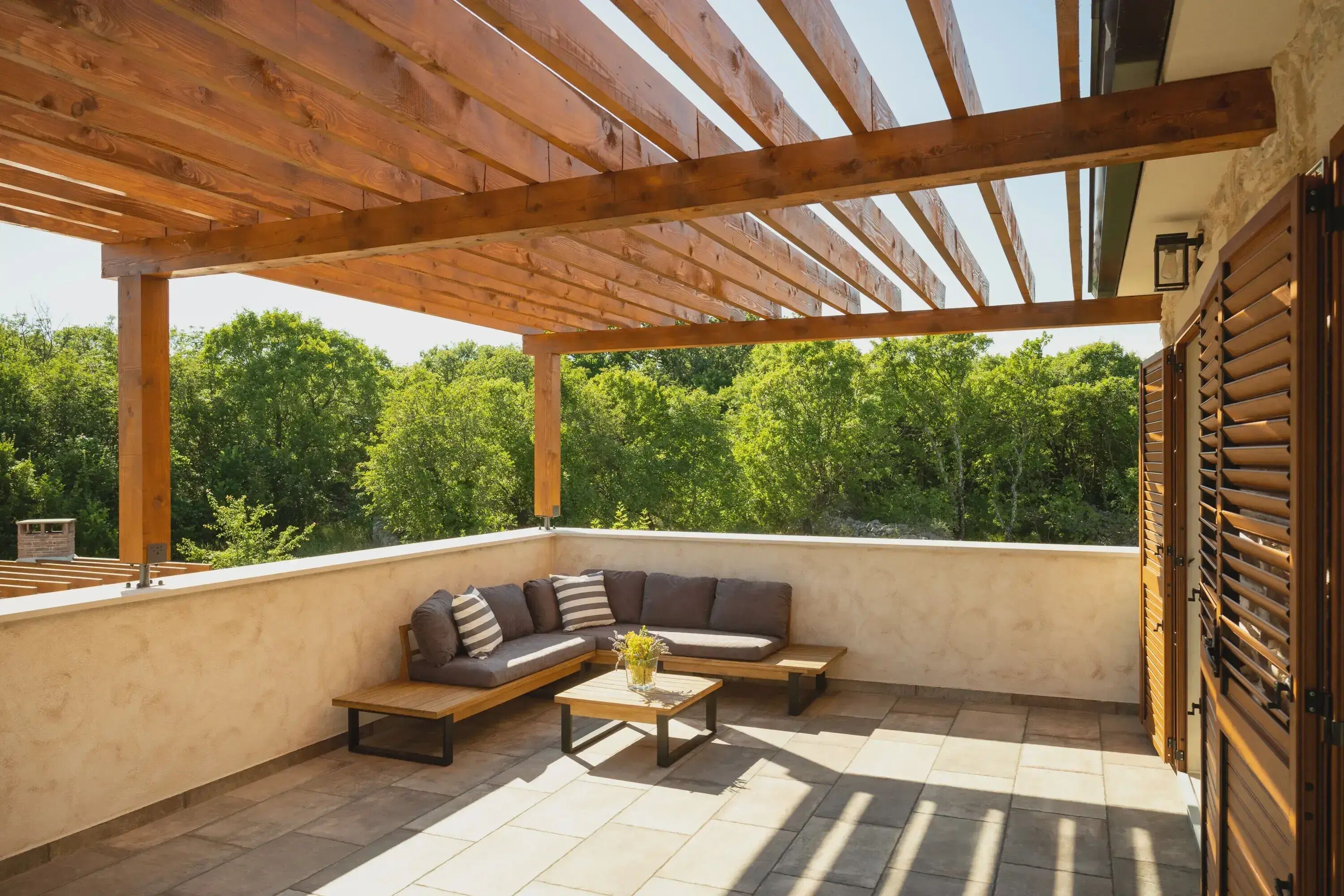 Un patio al aire libre con una pérgola de madera, un sofá modular, una mesita y vistas a zonas verdes al fondo.