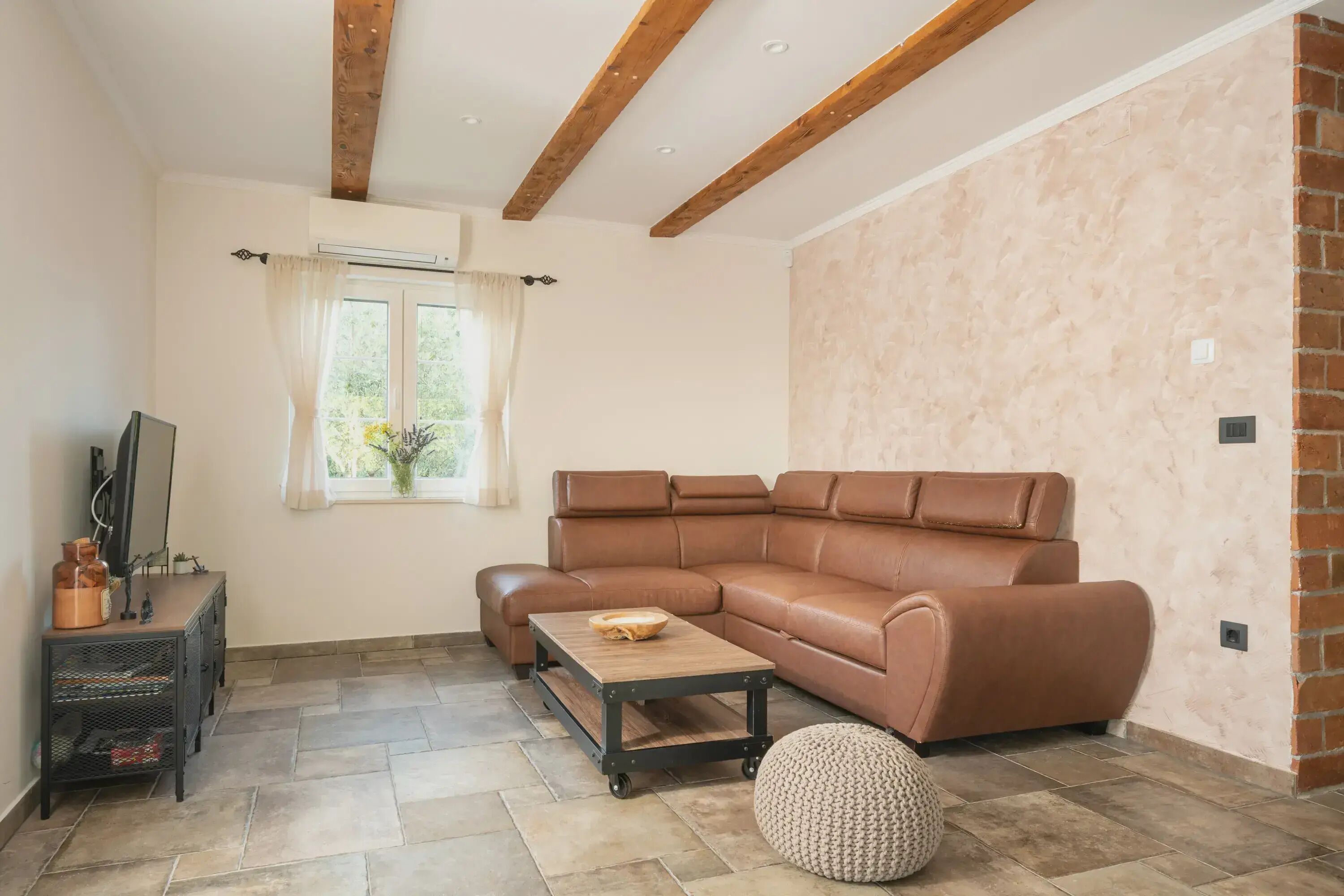 Una sala de estar con un sofá seccional de cuero marrón, una mesa de centro de madera, un mueble para televisión negro y una ventana con cortinas.