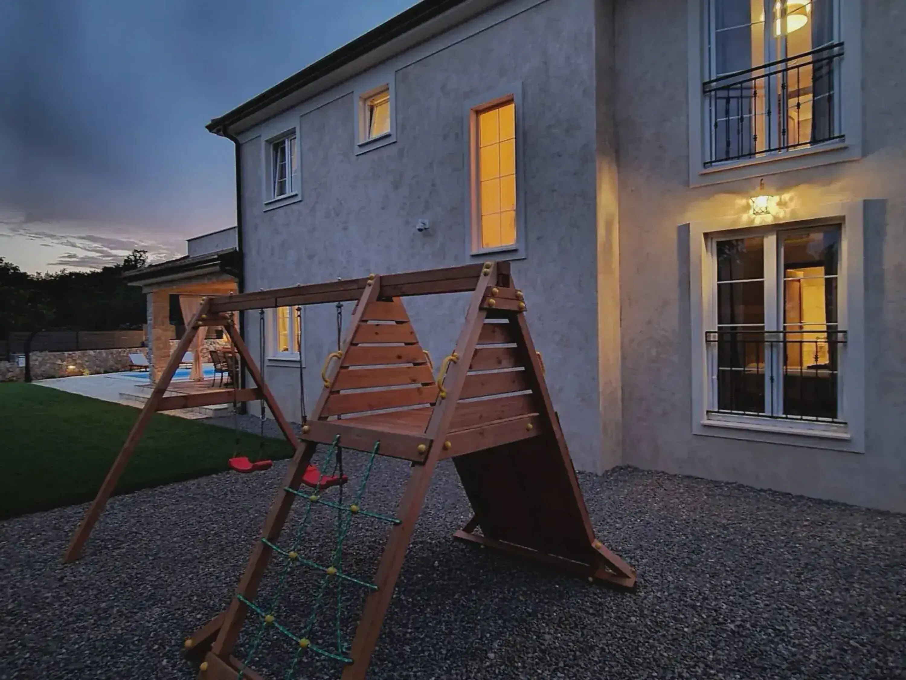 A wooden swing set with a net in front of a house with a pool in the background.