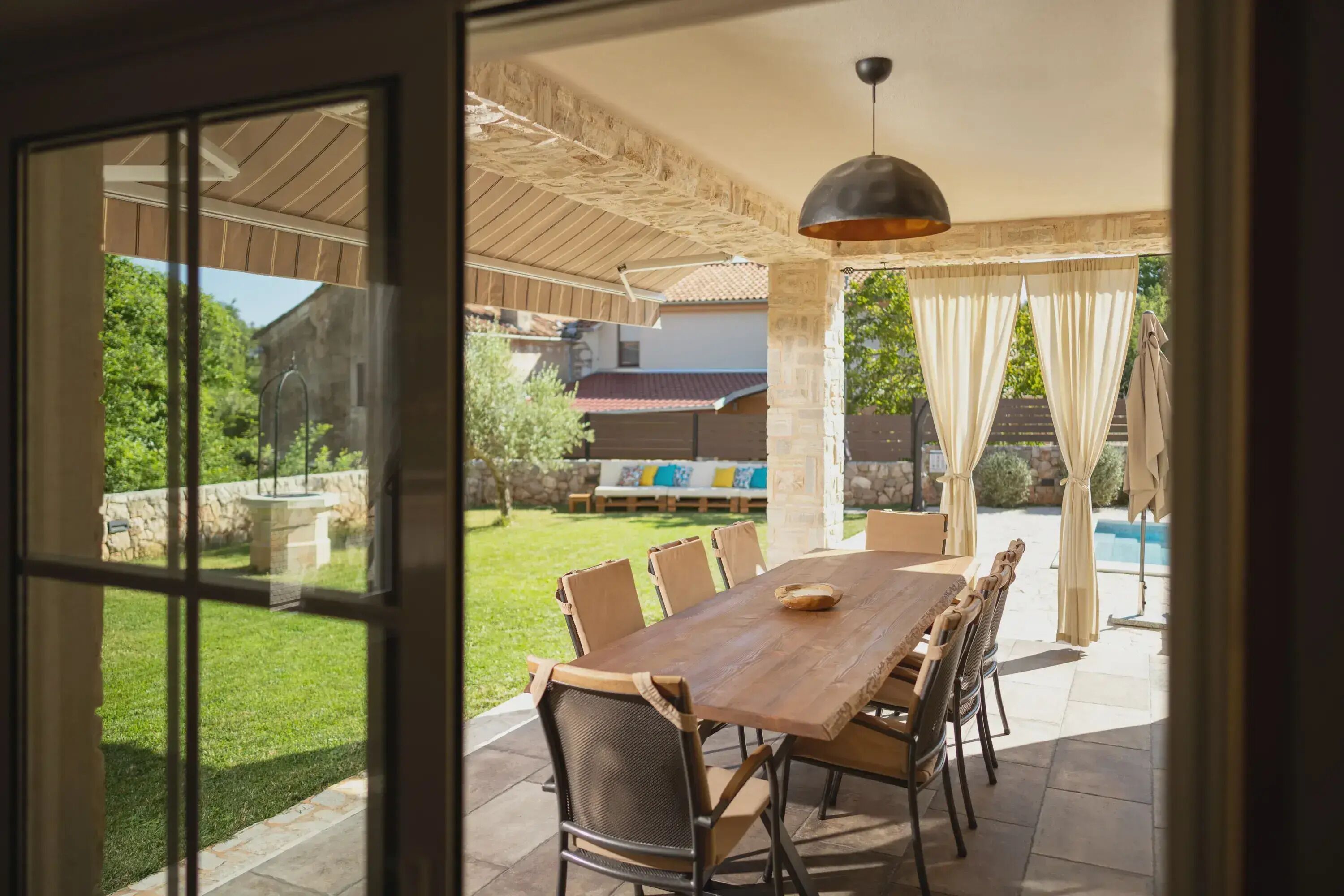 Eine Terrasse im Freien mit einem Holztisch und Stühlen, einer hängenden Leuchte sowie Blick auf Pool und Garten.