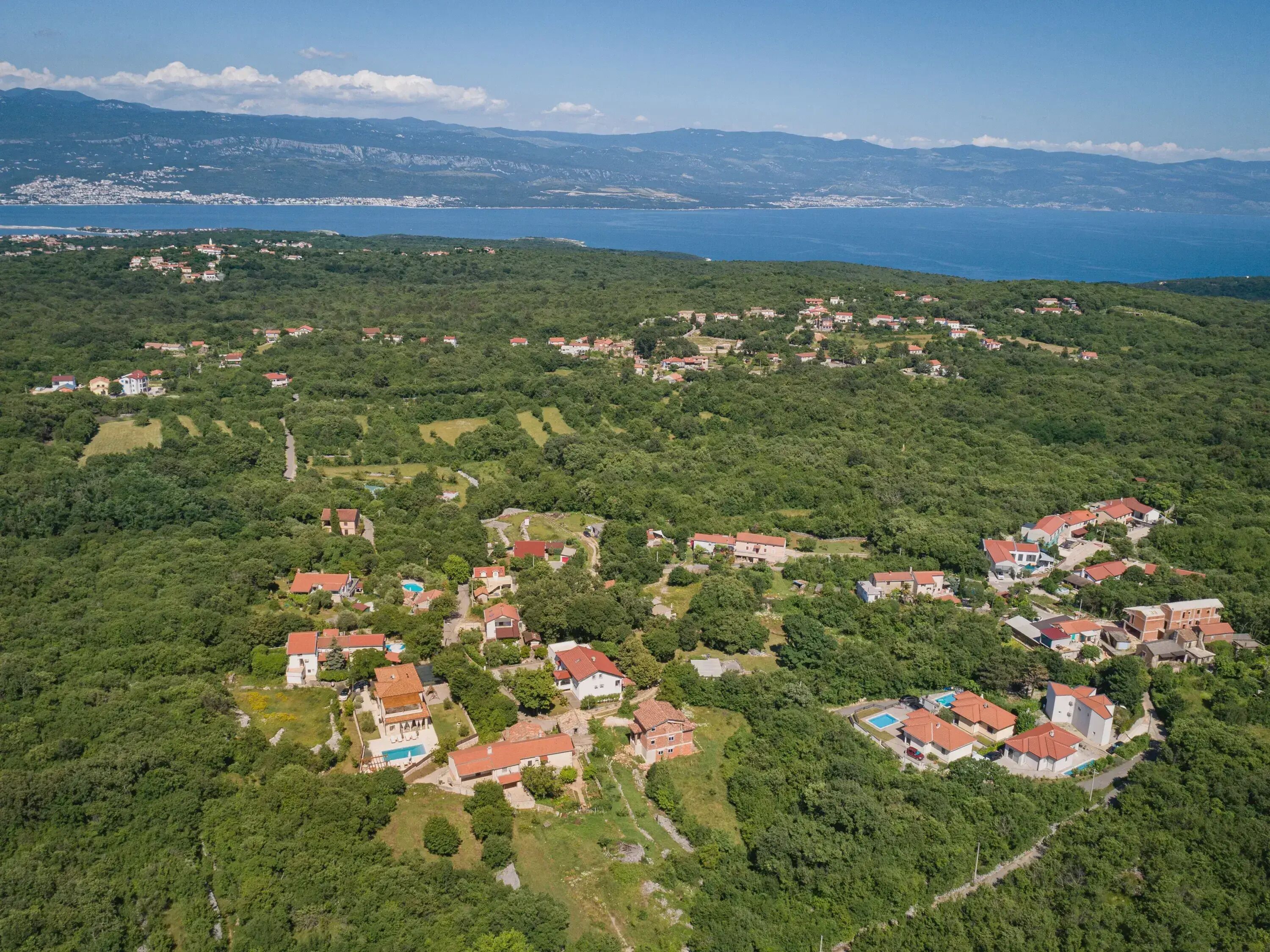 Vue aérienne d’un village perché sur une colline, avec ses maisons aux toits rouges, ses champs verdoyants et une côte visible à l’arrière-plan.