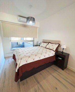 A bedroom with a bed, bedside tables, a window with blinds, an air conditioning unit, and a hanging light fixture.