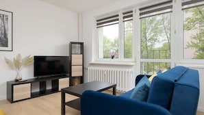 A modern living room with a large flat-screen TV, a blue sofa, a small coffee table, and a window with a view of trees.