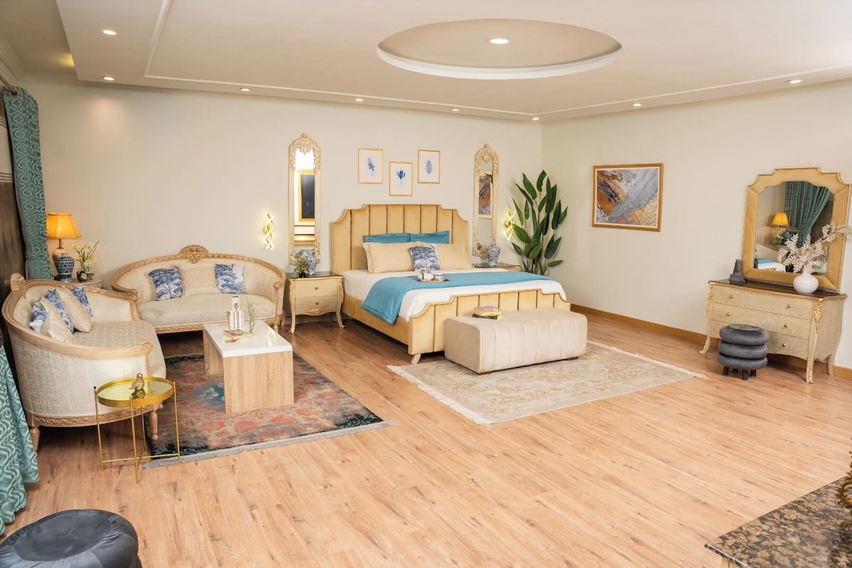 A spacious bedroom with a large bed, two armchairs, a small table, and a vanity with a mirror.