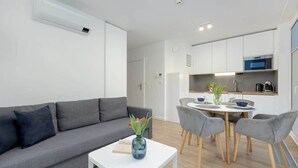Una sala de estar moderna con un sofá gris, una mesa de centro blanca con un jarrón, y un comedor con una mesa redonda y sillas grises.