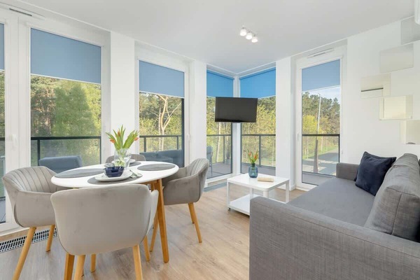 A bright living room with large windows, a round dining table, and modern decor. The room has light wood flooring and a cozy atmosphere.