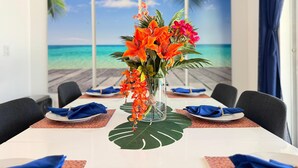 A dining table set with blue napkins, a glass vase with orange flowers, and a tropical beach view through a window. - 8BR Pool & Arcade – Enchanted Escape Retreat (Kissimmee)