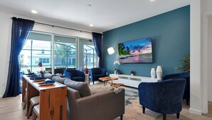 A modern living room with a sofa, chairs, a dining table, and a large window with curtains. - 8BR Pool & Arcade – Enchanted Escape Retreat (Kissimmee)