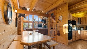 Interior rústico de una cabaña con una mesa de comedor de madera, bancos, una zona de cocina y un ventanal con vistas a un bosque.