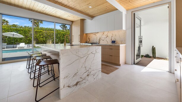 A modern kitchen with a marble island, wooden ceiling, and large windows overlooking a pool. - Desert Willow-Iconic Butterfly Alexander (Palm Springs)
