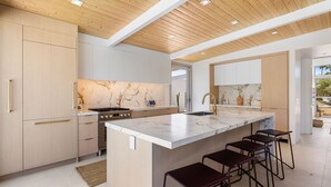 A modern kitchen with a marble backsplash, wooden ceiling, and a central island with bar stools. - Desert Willow-Iconic Butterfly Alexander (Palm Springs)