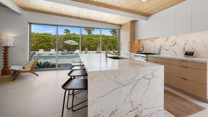A modern kitchen with a marble countertop, wooden cabinets, and a large window overlooking a pool and greenery. - Desert Willow-Iconic Butterfly Alexander (Palm Springs)