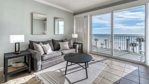 Un salon avec un canapé gris, deux tables d’appoint avec des lampes, une table basse ronde et une grande porte-fenêtre coulissante donnant sur un balcon avec vue sur la plage.