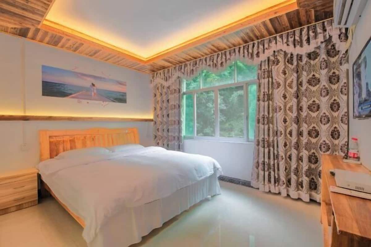Un dormitorio con cabecera de madera, cama con ropa de cama blanca, mesita de noche, ventana con cortinas estampadas y un cuadro en la pared.