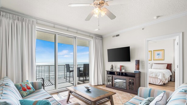 A spacious living room with a large sliding glass door leading to a balcony, a ceiling fan, a flat-screen TV, and a cozy seating area with striped cushions. - Big Beachfront Balcony for Parties of 7 with a set of beach chairs included! (Panama City Beach)