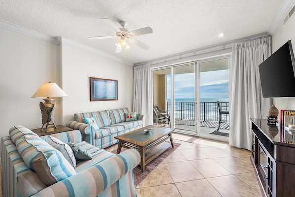 A living room with striped sofas, a coffee table, a ceiling fan, a TV, and a balcony with a view. - Big Beachfront Balcony for Parties of 7 with a set of beach chairs included! (Panama City Beach)