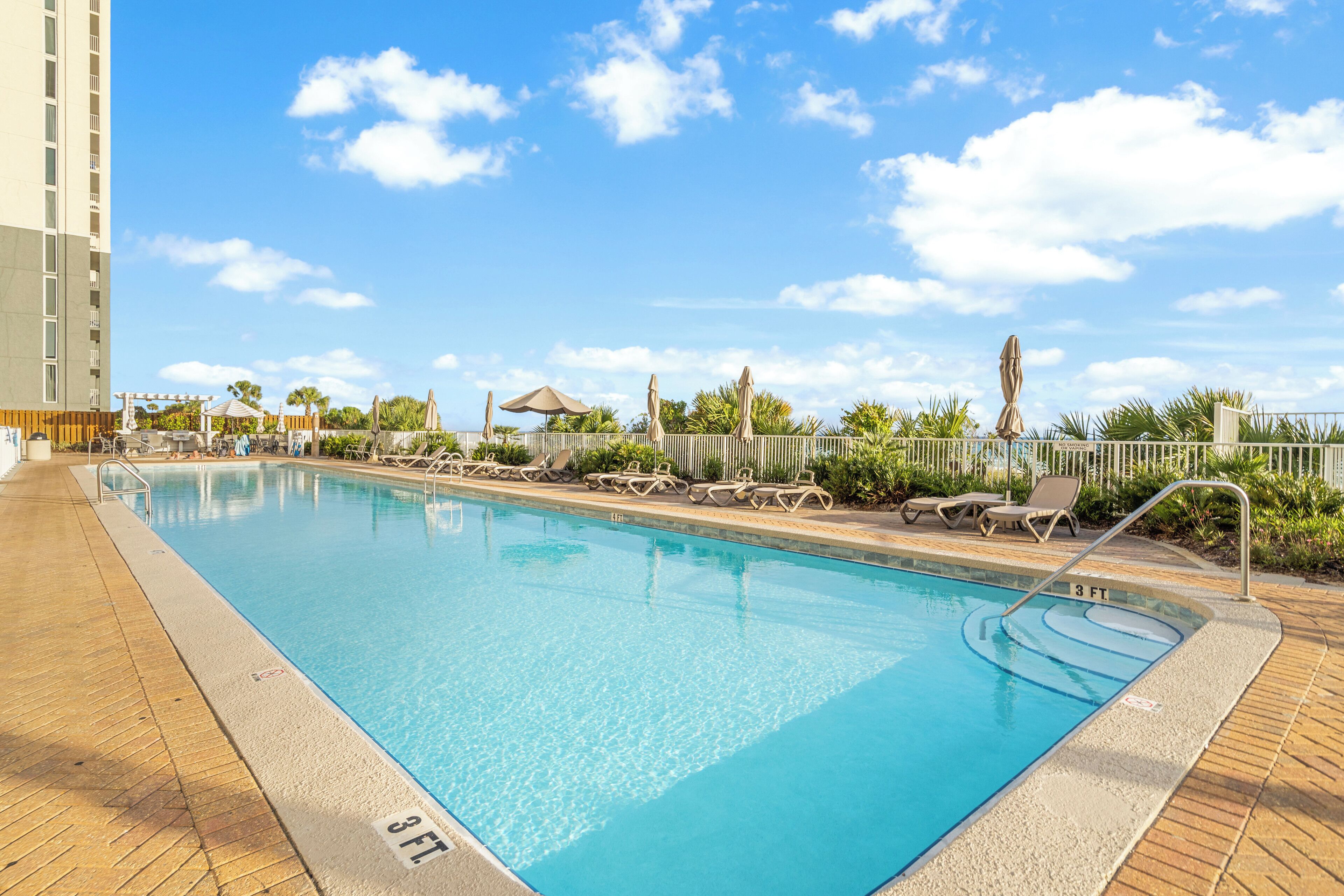 A swimming pool with lounge chairs and umbrellas, surrounded by a fence and greenery.