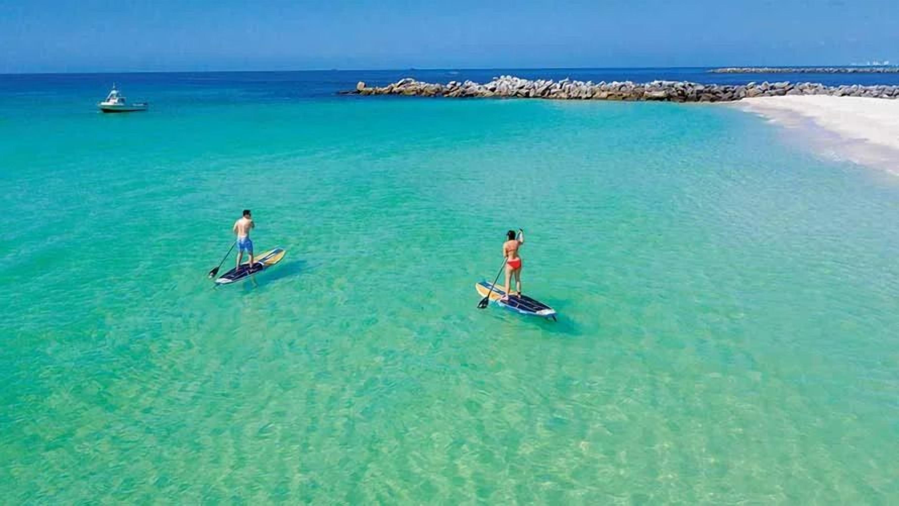 Two people paddleboarding in clear turquoise water with a boat in the background and a rocky shoreline.