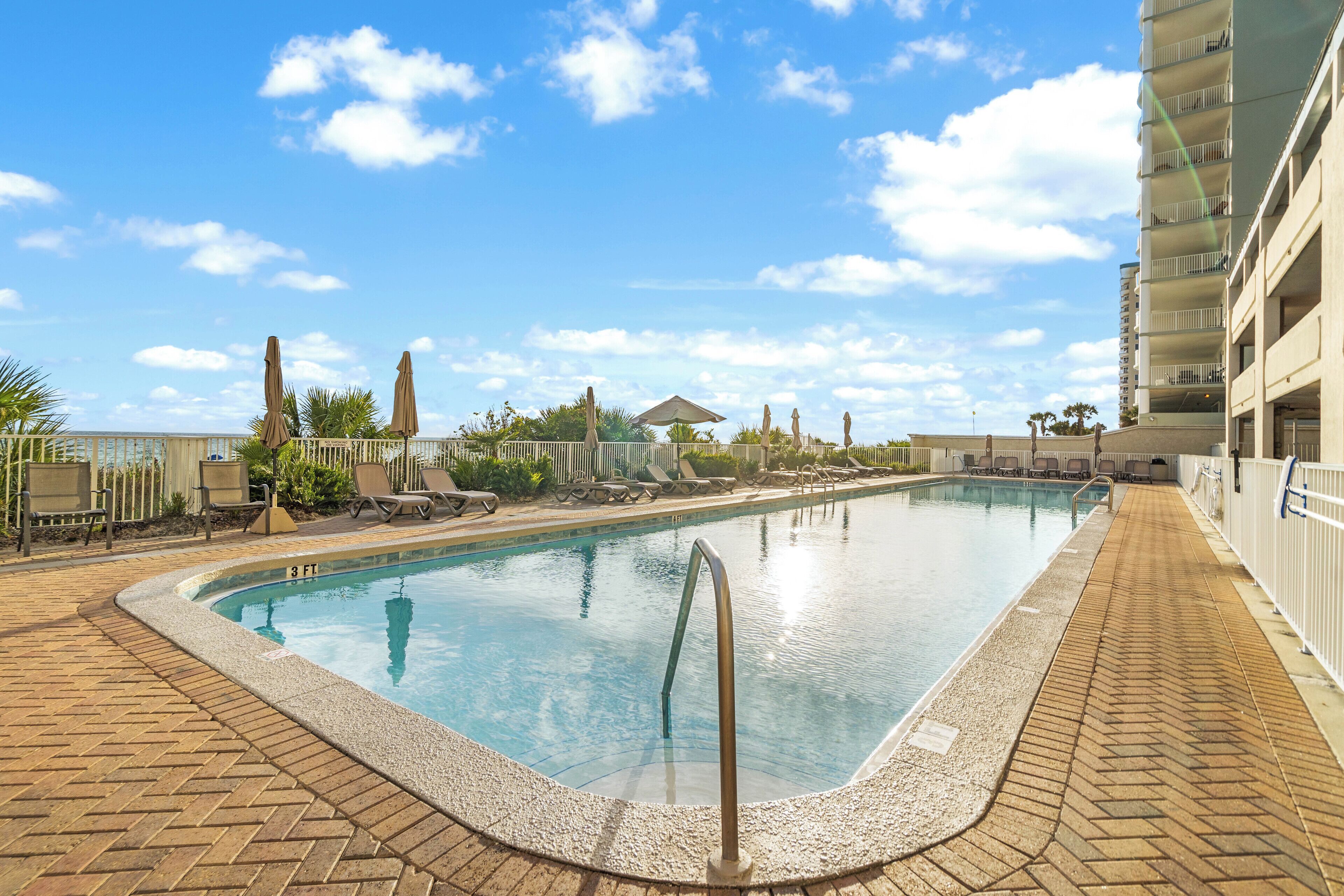 A rectangular swimming pool with a brick border, surrounded by lounge chairs and umbrellas, with a clear blue sky in the background.