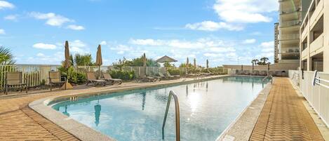 A rectangular swimming pool with a brick border, surrounded by lounge chairs and umbrellas, with a clear blue sky in the background.