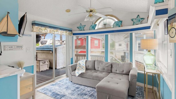 A room with a sofa, a ceiling fan, a window with blinds, and a small table with a lamp.