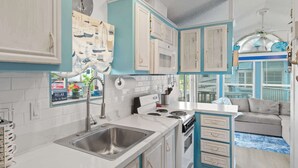 A kitchen with white and blue cabinets, a stainless steel sink, a stove, and a dining area with a sofa.