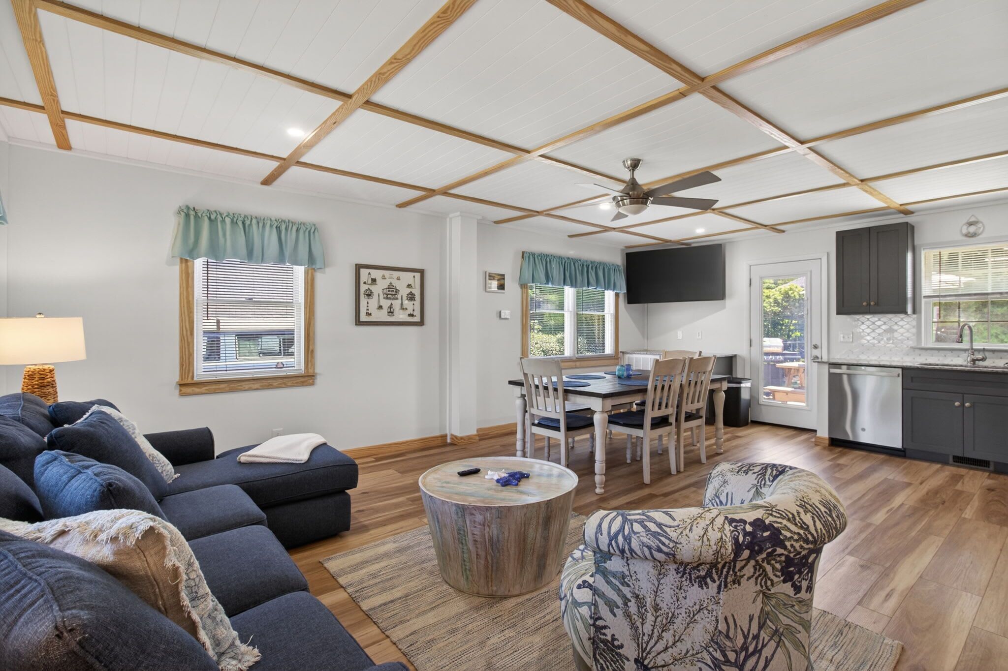 A living room with a sofa, a round coffee table, and a patterned armchair. There is a dining area with a table and chairs, and a kitchen with a sink and cabinets.