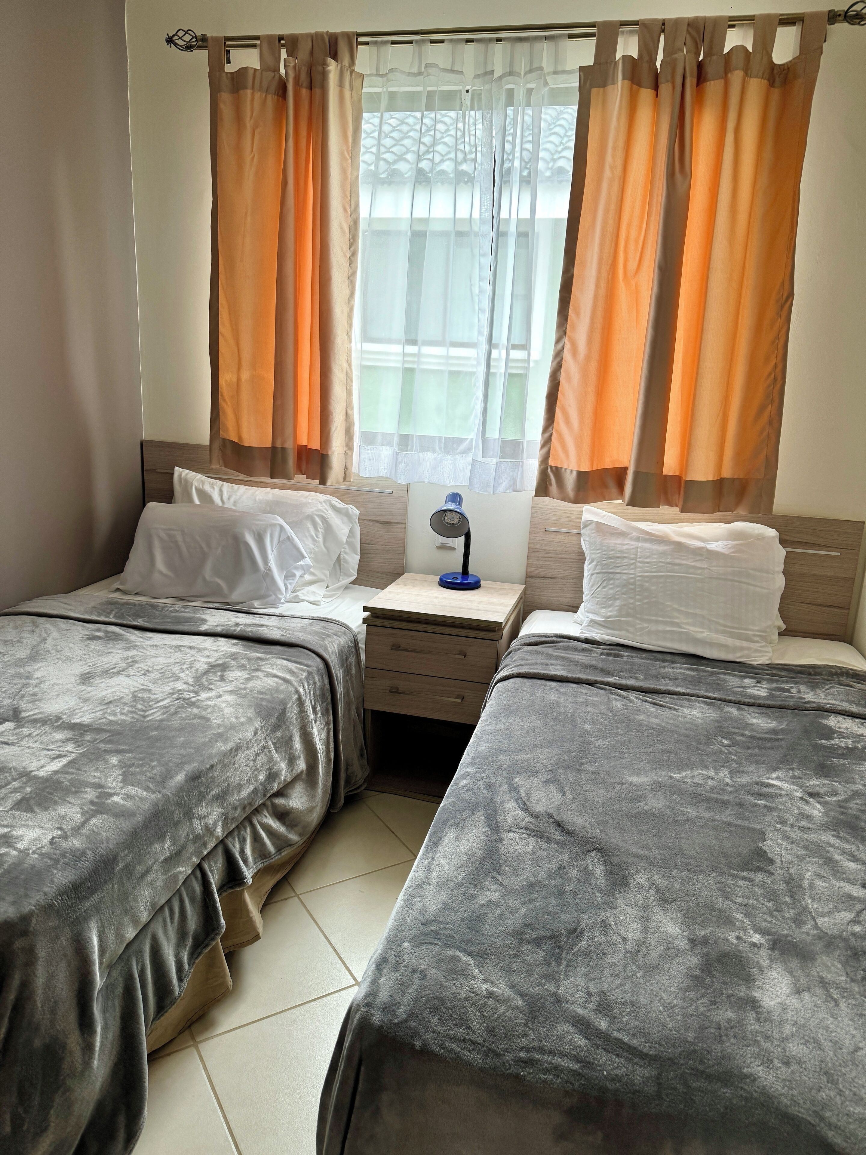 A hotel room with two beds, a nightstand with a lamp, and a window with orange curtains.