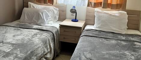A hotel room with two beds, a nightstand with a lamp, and a window with orange curtains.