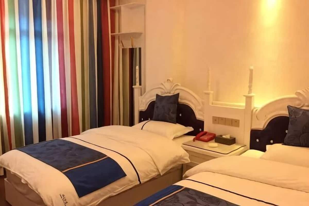 A hotel room with two beds, a bedside table with a lamp, and a window with vertical blinds.