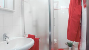Ein modernes Badezimmer mit Waschbecken in Weiß, roten Handtüchern, einer Glasdusche und einem kleinen Topfpflanzen.
