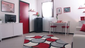 Ein modernes Wohnzimmer mit einem weißen Sofa, roten Kissen, einem Essbereich mit Tisch und Stühlen, einer roten Tür und einem großen Fenster mit Vorhängen.