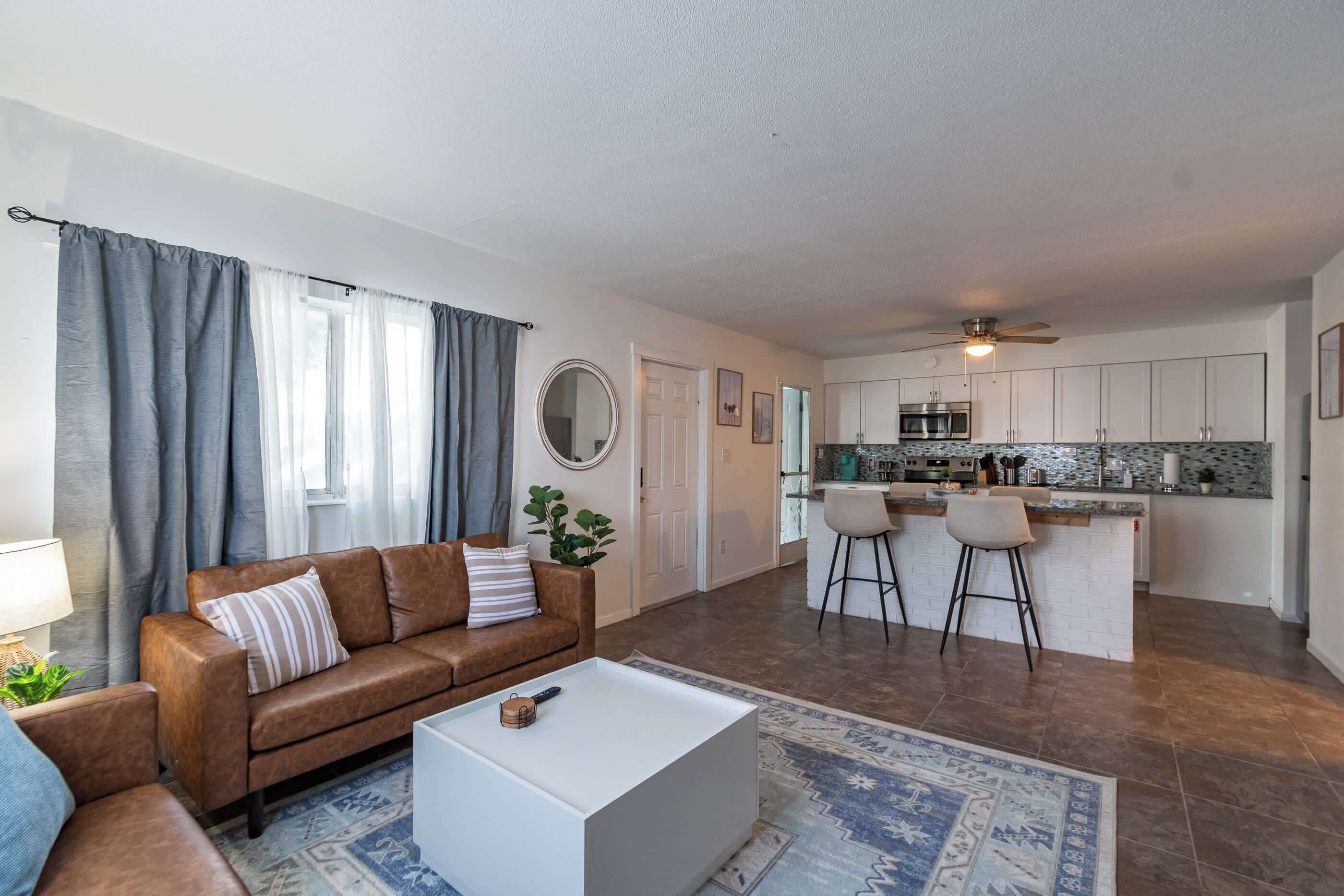 A modern living room with a brown leather sofa, a white coffee table, and a patterned rug. A kitchen with white cabinets and a central island is visible in the background.
