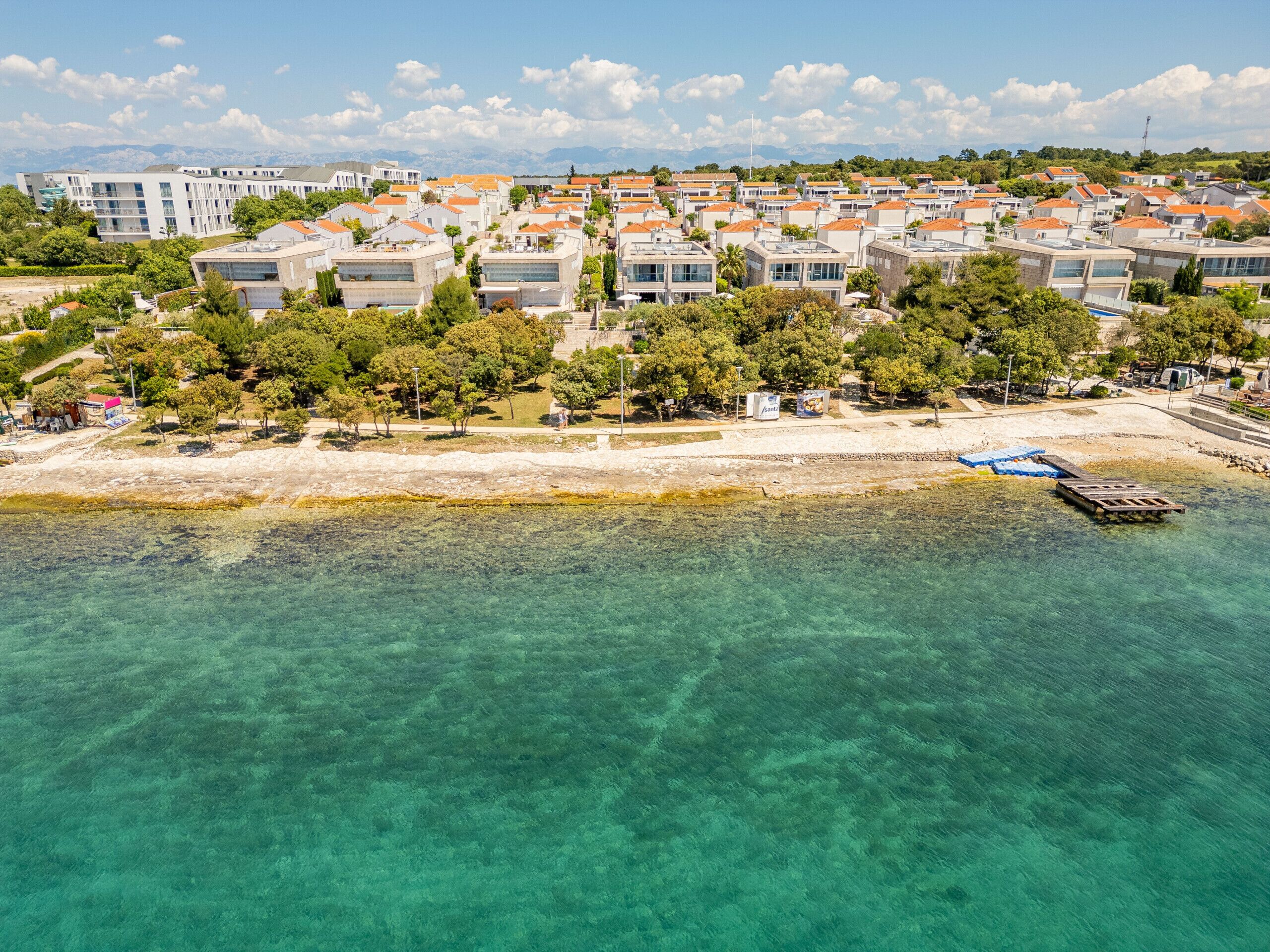 Vista aérea de una zona residencial costera con aguas turquesas cristalinas, una playa de arena y una hilera de edificios modernos con tejados anaranjados.
