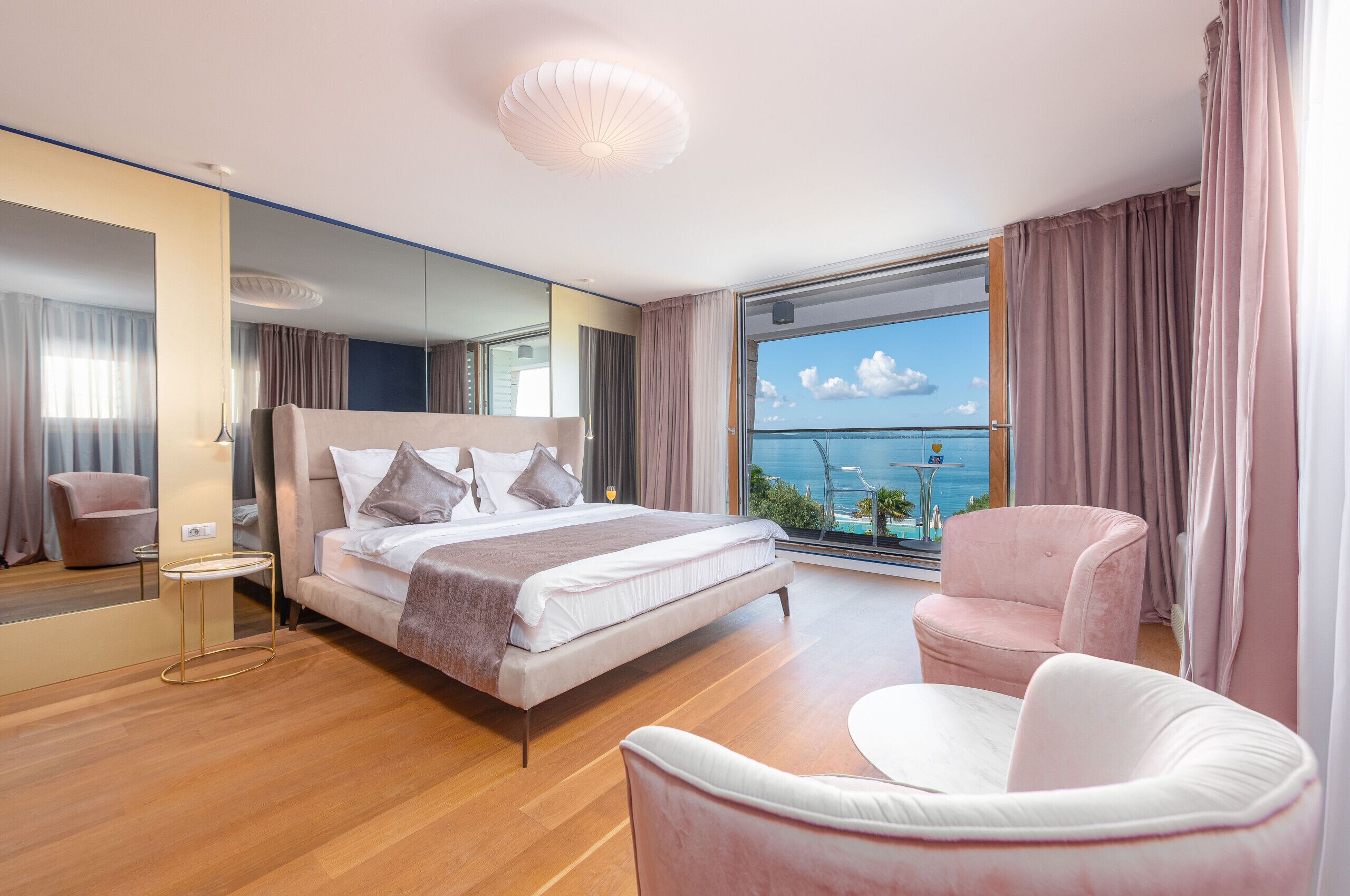 A modern hotel room with a large bed, a sitting area, and a balcony with a view of the sea.