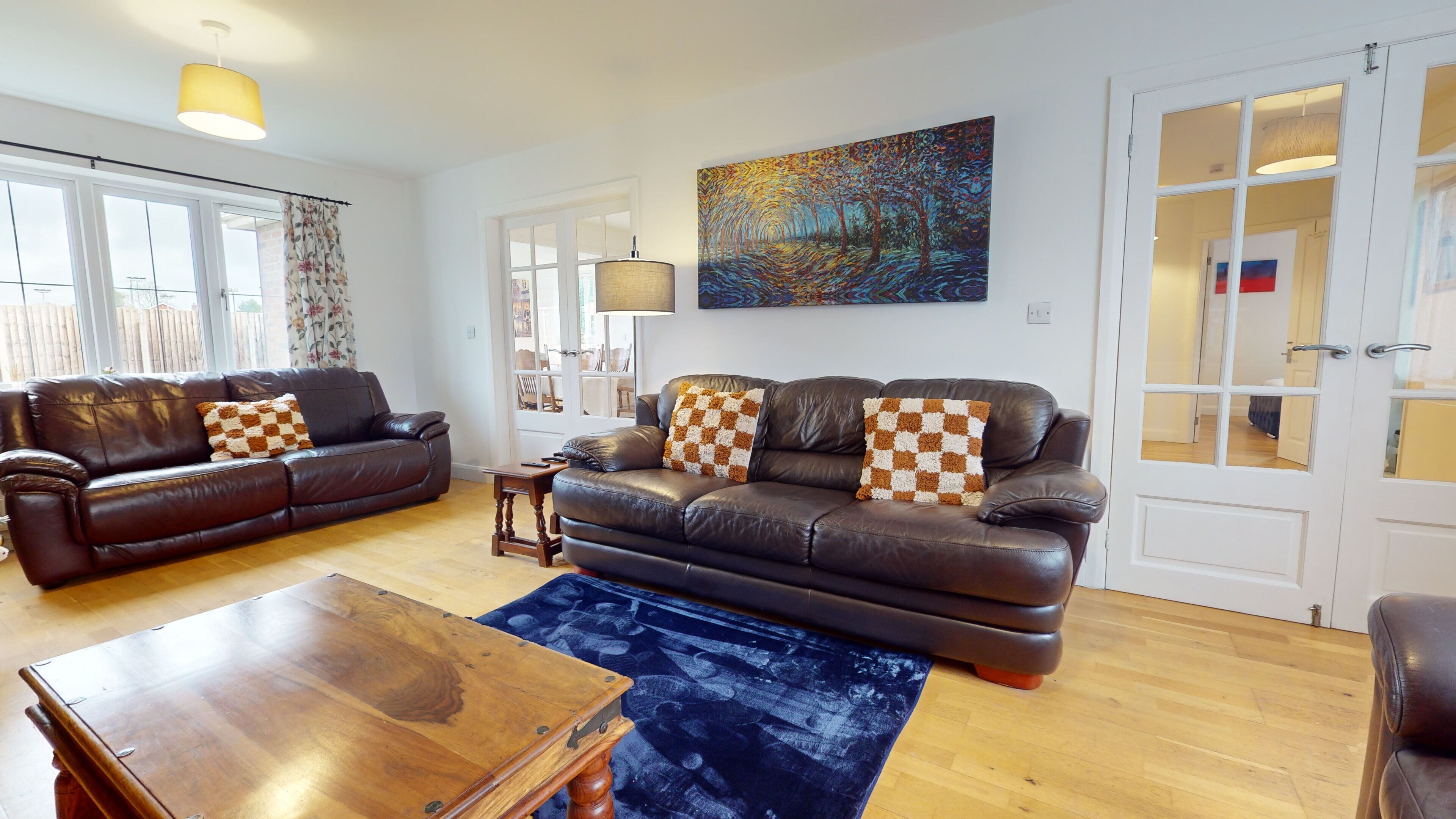 A living room with two leather sofas, a wooden coffee table, and a blue rug. There are large windows and a painting on the wall.