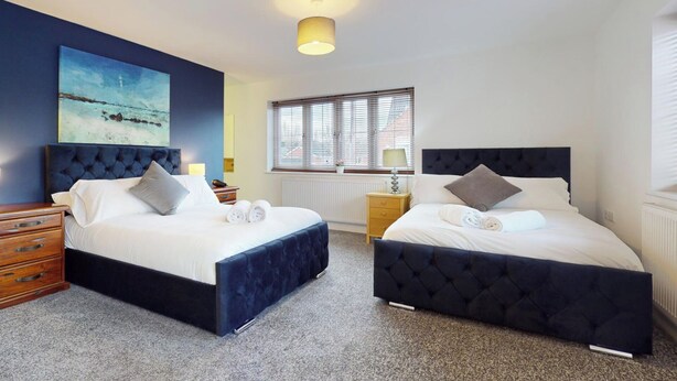 A hotel room with two beds, a large painting on the wall, a window with blinds, and a ceiling light. - 5-bedroom Home - Sleeps 16 - Ideal for Groups (Doncaster)
