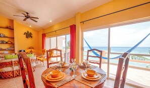 Un coin repas avec une table dressée pour quatre personnes, de grandes fenêtres offrant une vue sur l’océan et une bibliothèque remplie de livres.