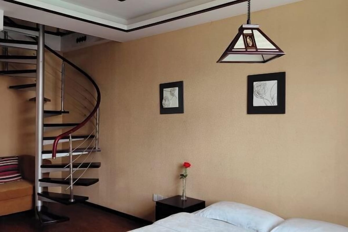 A bedroom with a bed, a nightstand, a vase with a single rose, a framed picture, a second framed picture, and a spiral staircase.
